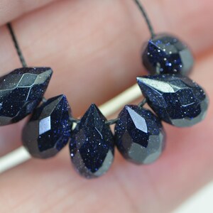 May include: A strand of dark blue gemstone beads, each bead is faceted and shaped like a teardrop. The beads are strung on a black cord.