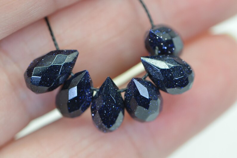 May include: A strand of dark blue gemstone beads, each bead is faceted and shaped like a teardrop. The beads are strung on a black cord.