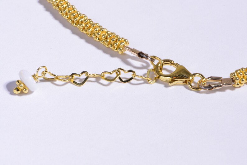 May include: Gold chain necklace with a white bead and heart-shaped links. The necklace has a lobster clasp closure.