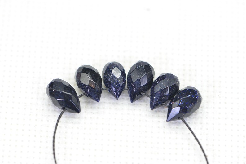 May include: A strand of dark blue gemstone beads with a faceted teardrop shape. The beads are strung on a black thread.