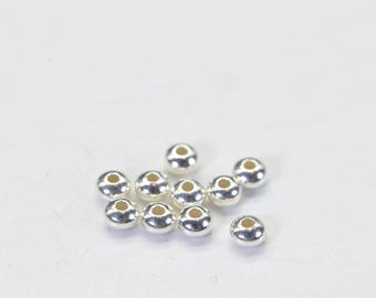 May include: A collection of small, shiny, silver-colored beads with a hole through the center. The beads are scattered on a white surface. These beads are likely used for jewelry making or other craft projects.