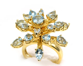 May include: A gold ring with a flower-shaped design featuring multiple light blue gemstones. The ring has a delicate, intricate design.