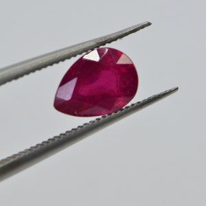 May include: A pear-shaped ruby gemstone held by tweezers. The ruby is a deep red color and has a smooth, polished surface.