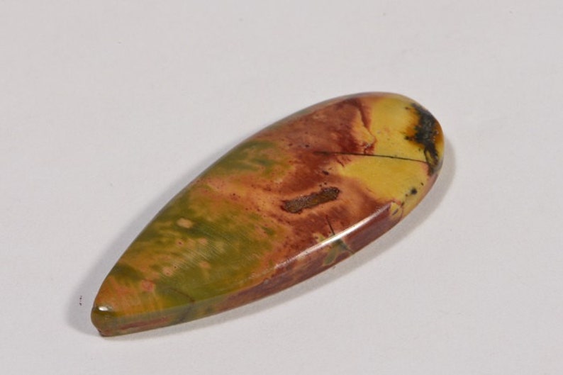 May include: A polished, teardrop-shaped gemstone with a mix of colors. The stone features a blend of green, yellow, and red hues, with darker brown and black accents. The surface is smooth and glossy.