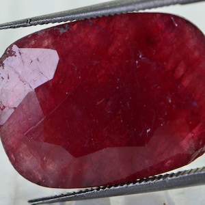 May include: A deep red gemstone with a faceted cut. The gemstone is oval shaped and has a smooth, polished surface.