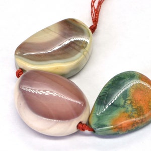 May include: Three smooth, polished stones with unique patterns and colors. The stones are strung together with red cord and are likely used for jewelry making.