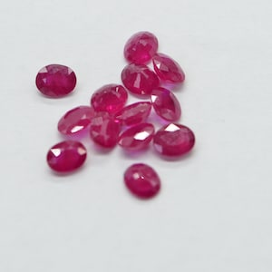 May include: A collection of faceted, oval-shaped, and round rubies in varying sizes. The gemstones are a deep, vibrant red color and are scattered on a white surface. The light catches the facets, creating a sparkling effect. Ideal for jewelry making.