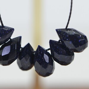 May include: A strand of dark blue gemstone beads, each bead is faceted and shaped like a teardrop. The beads are strung on a black cord.