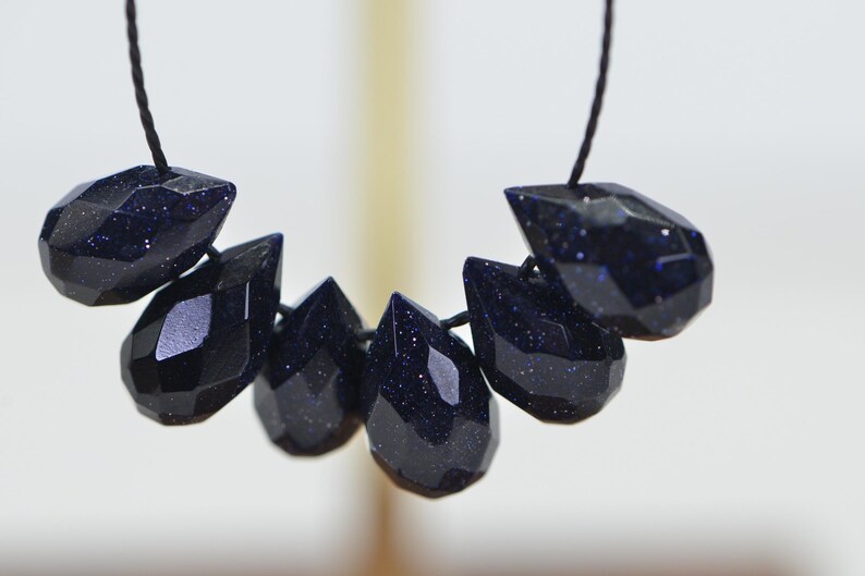May include: A strand of dark blue gemstone beads, each bead is faceted and shaped like a teardrop. The beads are strung on a black cord.