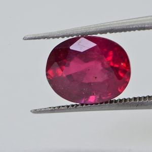 May include: A vibrant red oval-shaped gemstone held by tweezers. The gemstone has a faceted cut and is a deep red colour.
