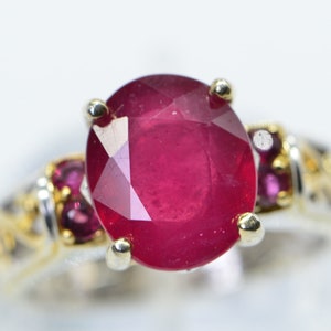 May include: A ring featuring a large oval ruby gemstone, surrounded by smaller ruby stones. The ring is crafted from gold and silver.