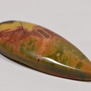 May include: A polished, elongated gemstone with a teardrop shape. The stone features a blend of colors, including olive green, mustard yellow, and reddish-brown hues. The surface has a smooth, glossy finish, and the stone appears to be a natural mineral.