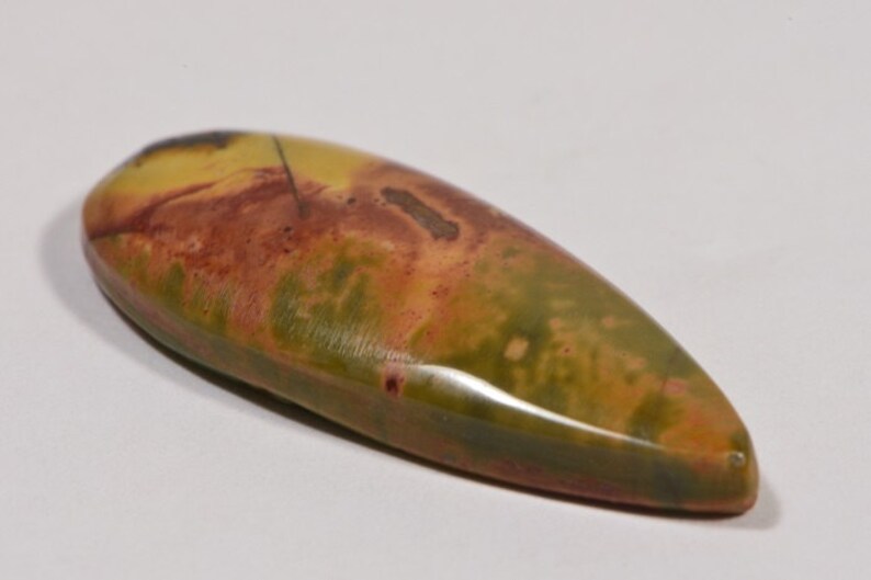 May include: A polished, elongated gemstone with a teardrop shape. The stone features a blend of colors, including olive green, mustard yellow, and reddish-brown hues. The surface has a smooth, glossy finish, and the stone appears to be a natural mineral.