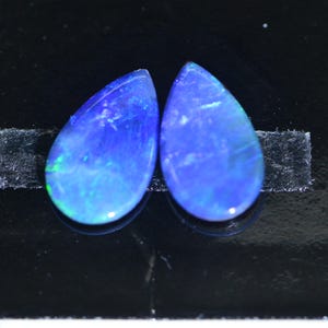 Puede incluir: Dos ópalos en forma de pera, con un vibrante color azul y juegos de color internos. Las piedras preciosas se presentan sobre un fondo oscuro, resaltando sus cualidades iridiscentes. Los ópalos tienen una superficie pulida y lisa.