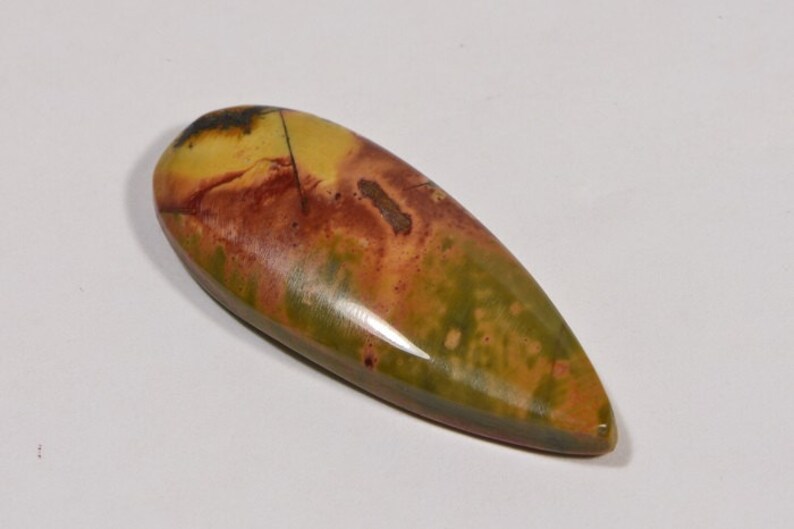 May include: A polished, teardrop-shaped gemstone with a blend of colors. The stone features a base of olive green, transitioning to shades of yellow and brown with reddish-brown accents. A dark, almost black, area is visible at the top.