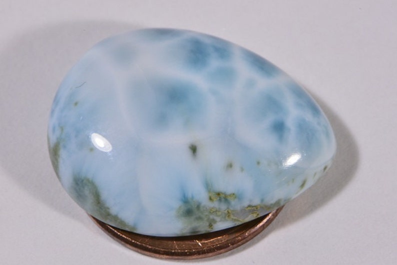 May include: A light blue cabochon gemstone with a white and blue marbled pattern. The gemstone is about the size of a US penny.