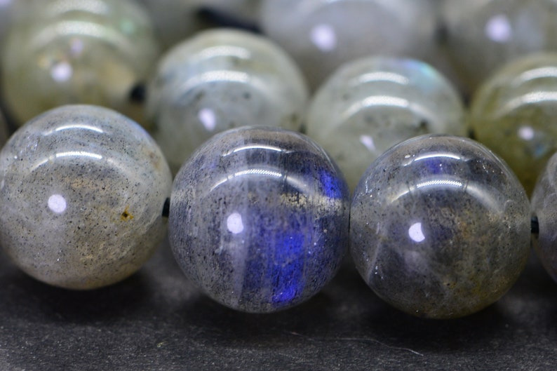 May include: A strand of round, grey labradorite beads with iridescent blue and green flashes. The beads are strung on a black cord.
