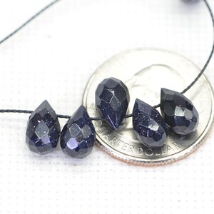 May include: Five dark blue teardrop-shaped beads with a sparkly finish, strung on a black cord. The beads are displayed next to a US quarter for size reference.