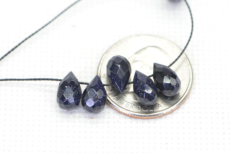 May include: Five dark blue teardrop-shaped beads with a sparkly finish, strung on a black cord. The beads are displayed next to a US quarter for size reference.