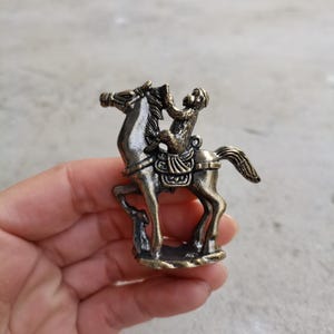 May include: A bronze-colored figurine of a monkey riding a horse. The monkey is seated on a saddle, and the horse is in a trotting pose. The figurine has intricate details and a vintage aesthetic.