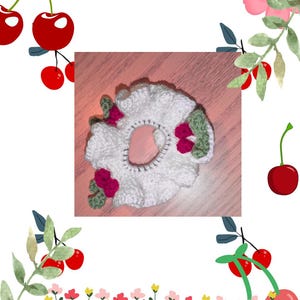 Könnte beinhalten: Weißes gehäkeltes Haargummi mit gerüschtem Rand, verziert mit grünen Blättern und rosa Blumenakzenten. Das Haargummi ist von Kirsch- und Blumenillustrationen umgeben, was eine skurrile, feminine Ästhetik erzeugt.