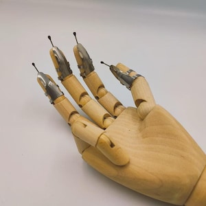 May include: A wooden hand model with silver-colored finger armor and small black spikes extending from the fingertips. The hand is posed against a white background, showcasing the detail of the articulated joints.