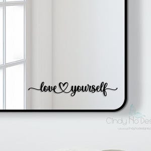 May include: Rectangular mirror with rounded corners and a black frame. The mirror reflects a bright window and the words "love yourself" in a cursive font. The mirror is mounted on a white wall.