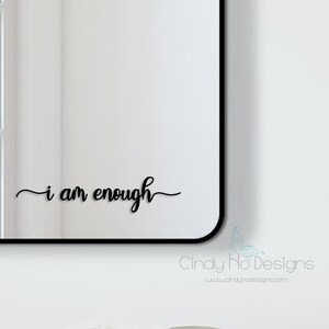 Pode incluir: Um espelho retangular com moldura preta e fundo branco que se reflete no vidro. O espelho tem o texto "i am enough" escrito em itálico na parte inferior.