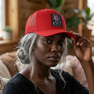May include: A bright red trucker hat featuring a mesh back and a rectangular patch on the front. The patch displays vertical blue and white stripes with a central red heart icon and the text "CERTIFIED GANGSTER OF LOVE" printed in white.