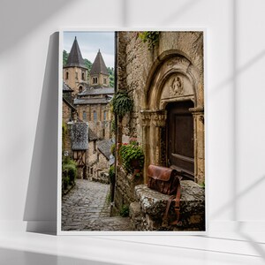 May include: A framed vertical print depicting a narrow cobblestone street in a historic village with stone buildings, a dark wooden arched doorway, and a brown leather satchel resting on a stone ledge in the foreground under green plants.