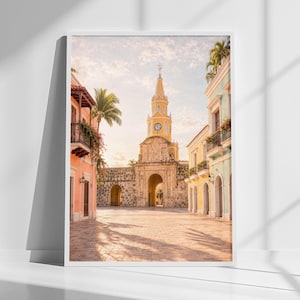 May include: A framed wall art print depicting a cobblestone plaza in Cartagena, Colombia. The scene features the iconic yellow Clock Tower gate, surrounded by colorful colonial buildings in shades of peach, yellow, and light blue under a bright sky.