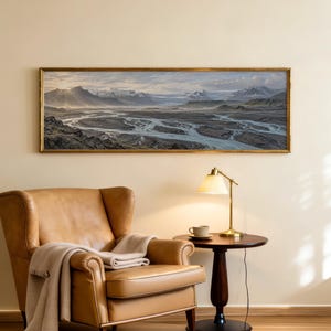 May include: A panoramic landscape painting in a gold frame depicting a glacial river valley with snow-capped mountains under a soft sky. Below, a tan leather wingback chair with a draped beige throw blanket sits next to a dark wood side table.