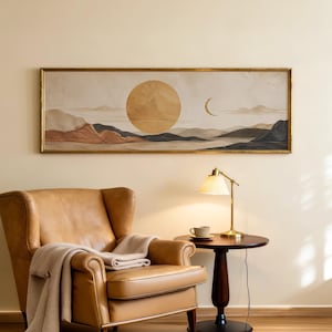 May include: A rectangular landscape wall art piece in a gold frame featuring a large golden sun and a crescent moon above layered mountains in shades of beige, brown, and dark gray, displayed horizontally on a plain wall above a leather armchair.