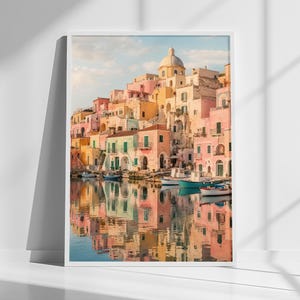 May include: A framed vertical wall art print depicting a coastal town with pastel-colored buildings in shades of pink, yellow, and peach, featuring a central domed structure, small boats floating on calm water, and clear reflections on the surface.