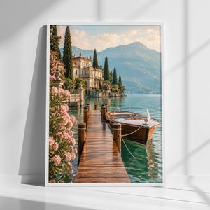Könnte beinhalten: Ein gerahmter Wandkunstdruck, der einen Holzsteg zeigt, der in türkisfarbenes Wasser ragt, mit einem polierten Holzmotorboot an der Seite. Eine große Villa mit Zypressen und rosa Oleanderblüten steht am Ufer unter Bergen.