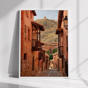 May include: A vertical wall art print depicting a narrow cobblestone street lined with tall, terracotta-colored buildings featuring wooden balconies. In the background, a rocky hill rises with a stone fortress wall visible against a clear sky.