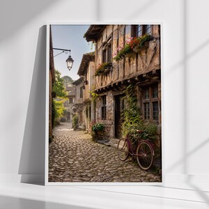 May include: A framed wall art print depicting a narrow cobblestone street in a historic village. The scene features timber-framed buildings with flower boxes, a vintage bicycle leaning against a stone wall, and a hanging black metal street lamp.