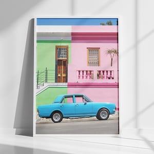 Può includere: Una stampa incorniciata con una scena di strada colorata. L'immagine mostra un'auto azzurra parcheggiata di fronte a un edificio dipinto di verde e rosa. Il cielo è blu. La stampa è esposta contro un muro bianco.
