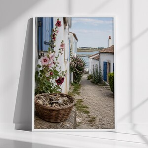 May include: A framed wall art print depicting a cobblestone path leading toward a coastal inlet. A woven basket filled with oyster shells sits in the foreground next to tall pink hollyhock flowers against a white building with blue shutters.