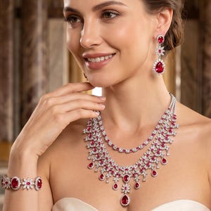 May include: A jewelry set consisting of a multi-layered necklace, drop earrings, and a bracelet, all featuring pear-shaped and round red gemstones surrounded by clear white stones set in a silver-toned metal frame with a polished finish.