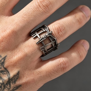 May include: A wide silver-toned metal ring featuring an openwork grid design that resembles a cage. The band is adorned with a central figure of a person with outstretched arms, integrated into the vertical and horizontal bars of the structure.