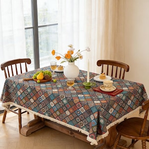 Könnte beinhalten: Ein Holztisch mit einer gemusterten Tischdecke in Blau-, Braun- und Orangetönen mit Fransen. Auf dem Tisch stehen eine Vase mit Blumen, Obst und Geschirr. Ein Kerze und Gläser sind ebenfalls vorhanden.