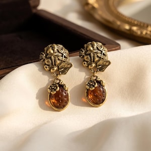 May include: A pair of drop earrings featuring a brass-toned metal top with a sculpted floral and leaf design, connected to a dangling, translucent amber-colored oval stone with a textured surface and a small metal flower accent at the top.