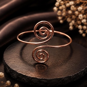Op de afbeelding: Een roségoudkleurige metalen armband met twee symmetrische, spiraalvormige uiteinden. De draadvormige band is gepolijst en glad en vormt een open, cirkelvormig ontwerp dat bij elk uiteinde in strakke spiralen krult.