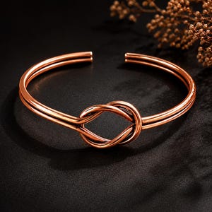 Op de afbeelding: Een gepolijste koperkleurige armband met een dubbelbandig ontwerp dat samenkomt in een centraal knoopdetail. De open armband heeft een gladde, reflecterende metalen afwerking en een cirkelvormig ontwerp met een strakke uitstraling.