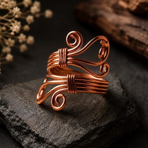 May include: A copper wire ring featuring a bypass design with two open ends. The metal wire is coiled into decorative spirals at each terminal and secured with smaller wire wraps along the band, creating a layered, open-ended structure.