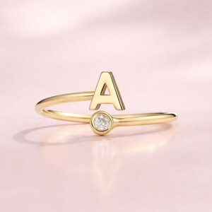 Peut inclure: Une bague dorée avec la lettre "A" et une petite pierre précieuse claire. L'anneau de la bague s'incurve pour rencontrer la lettre, créant un design délicat et élégant. La bague est posée sur un fond rose clair.