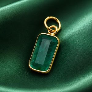 May include: A rectangular emerald pendant with a gold-colored frame and bail. The pendant is set against a backdrop of deep green satin fabric, creating a luxurious and elegant aesthetic. The gemstone is a vibrant green color.