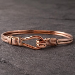 May include: A rose gold bracelet with a hook and eye clasp. The bracelet features a double band design with wire wrapping around the clasp and the ends of the bracelet. The jewelry is displayed on a dark gray surface.