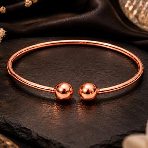 May include: A rose gold-toned metal cuff bracelet featuring a smooth, circular band that terminates in two polished spherical ball ends. The open-ended design is crafted from a reflective, metallic material with a warm, pinkish-copper hue.